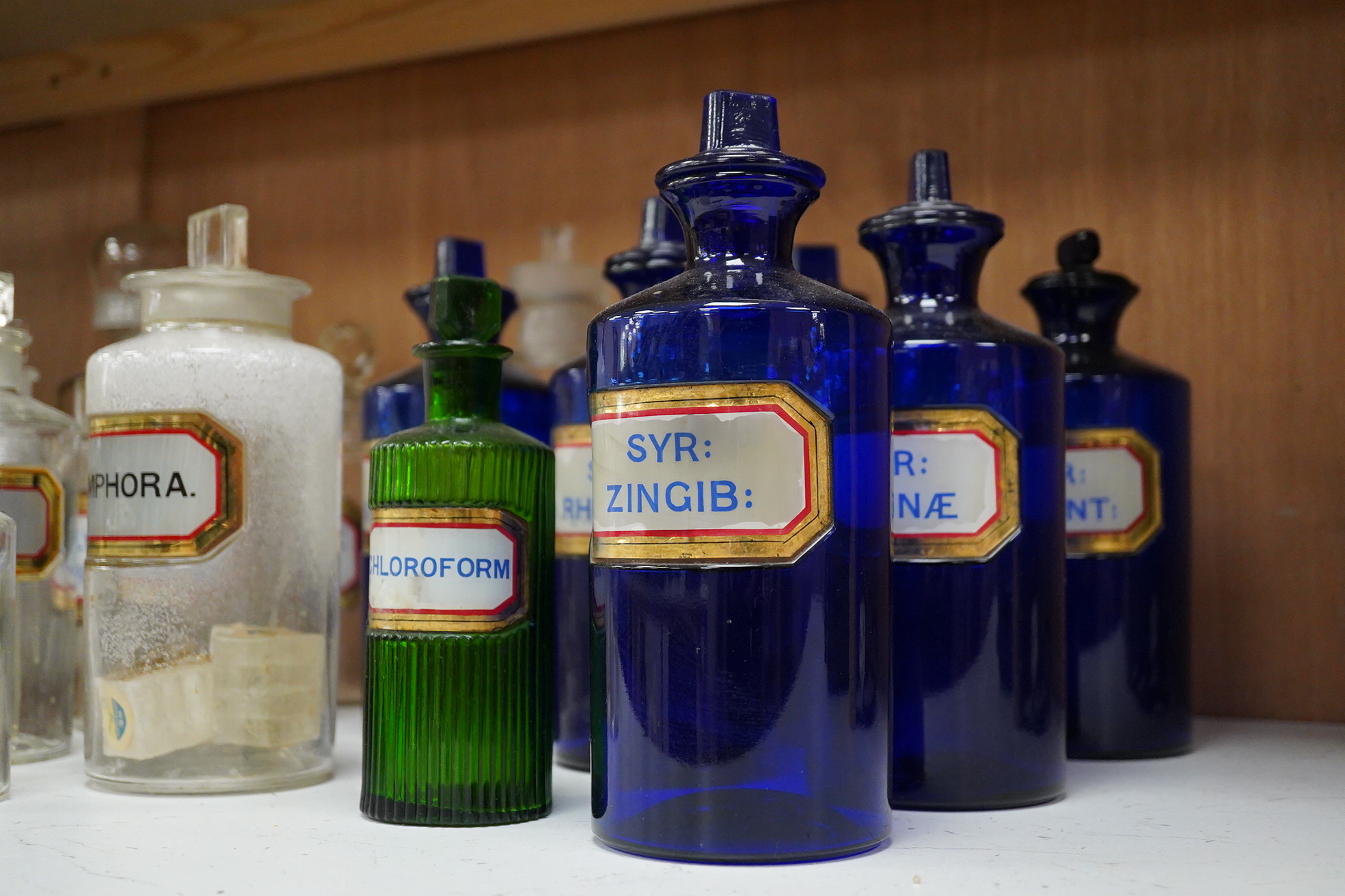Seventeen 19th/early 20th century glass apothecary jars with gilt rimmed labels; six blue glass, ten clear glass and a green glass example, labels including; Syr: Zingib:, Chloroform, Syr: Rhei:, Camphora, etc. tallest 2